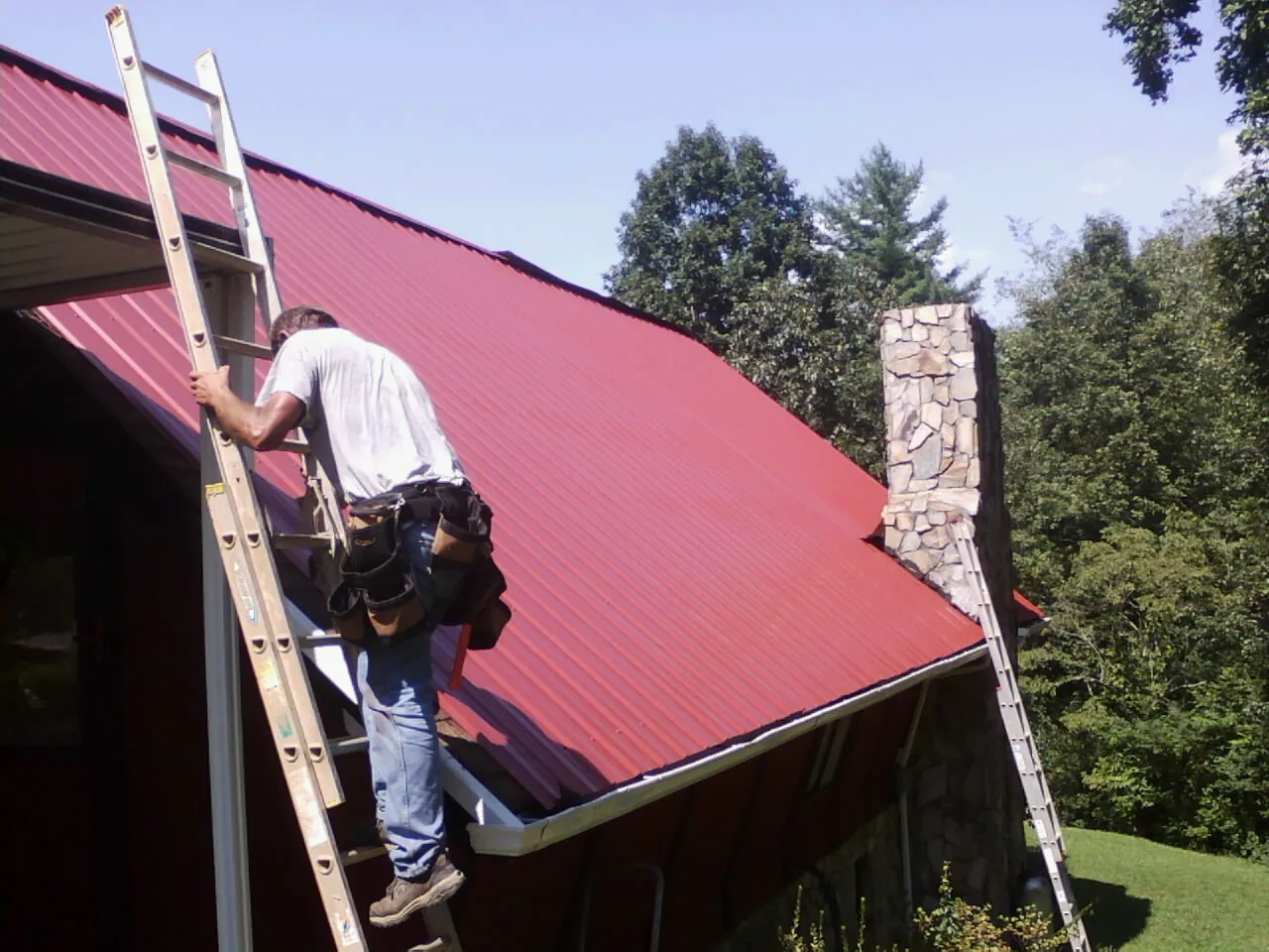 Roofer on a ladder beside a red metal roof and stone chimney — Soffit & Fascia Repair by Roofing Brothers in Rumsey